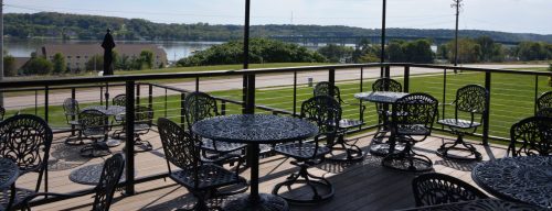 A scenic outdoor patio with black metal tables and chairs overlooking a river and green landscape.