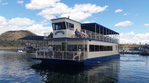 A large houseboat with multiple decks floats on calm water, surrounded by hills and a blue sky with clouds.