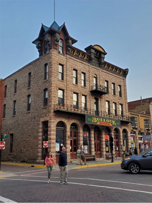 Historic stone building with ornate architecture, featuring a sign for "Bullock Hotel," with pedestrians and cars nearby.