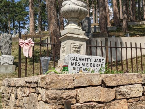 Gravestone of Calamity Jane, marked with a pink bow, surrounded by a stone fence in a wooded cemetery.