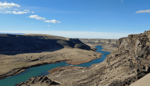 A scenic view of a winding river surrounded by rocky cliffs and open grassland under a clear blue sky.