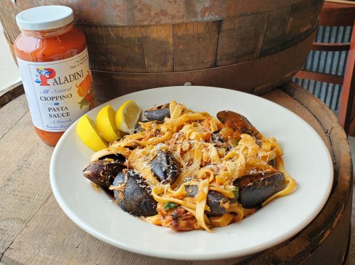 A plate of pasta with mussels, garnished with lemon and cheese, next to a jar of pasta sauce on a wooden barrel.