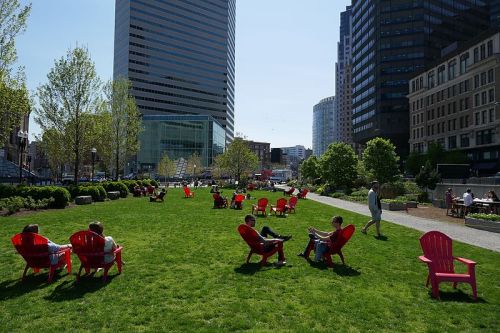 People relax in red chairs on a green lawn surrounded by tall buildings on a sunny day.