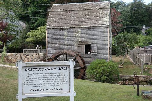 Historic Dexter's Grist Mill with a water wheel, surrounded by greenery and a sign detailing its restoration in 1961.