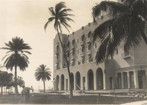 Black and white photo of a building with arched windows, surrounded by palm trees and a landscaped area.