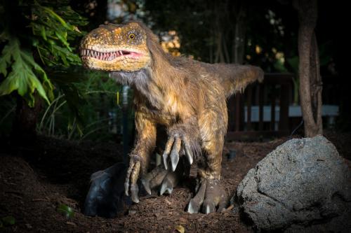 A lifelike dinosaur sculpture stands among trees and rocks, showcasing detailed features and a fierce expression.