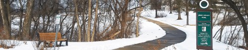 A snowy park path with a bench and a trail sign, surrounded by bare trees and a river in the background.