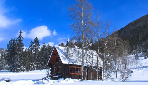 A cozy cabin surrounded by snow-covered trees and mountains under a bright blue sky.