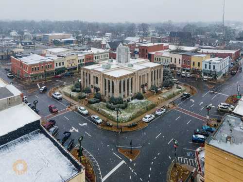 Aerial view of a snowy town square featuring a historic building, surrounded by shops and cars on a winter day.