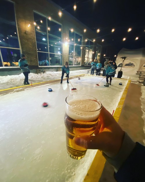 A hand holds a beer in the foreground, with people curling on an outdoor ice rink at night, string lights overhead.