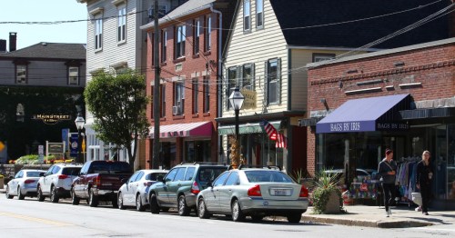 A quaint street lined with shops and parked cars, featuring a mix of brick and wooden buildings under clear blue skies.