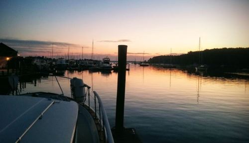 A serene sunset over a marina, with boats reflected in calm water and silhouettes of masts against the colorful sky.
