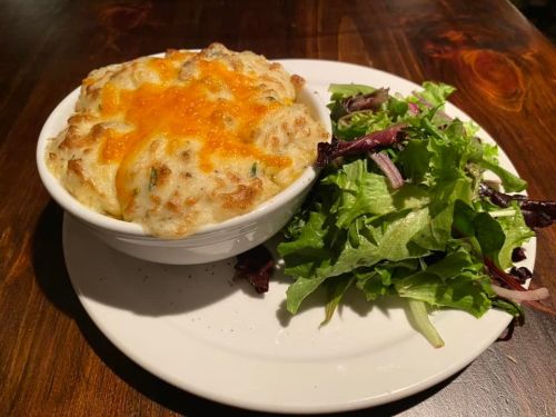 A bowl of cheesy baked pasta with a golden crust, served alongside a fresh green salad on a white plate.