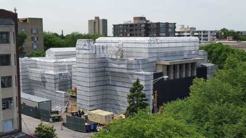 A large building wrapped in protective sheeting, surrounded by trees and construction equipment.