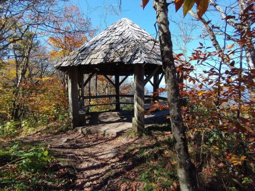 A wooden gazebo with a thatched roof surrounded by colorful autumn trees and a clear blue sky.
