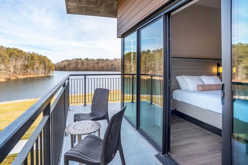 A balcony with two chairs overlooks a serene lake and forest, with a cozy bedroom visible through glass doors.