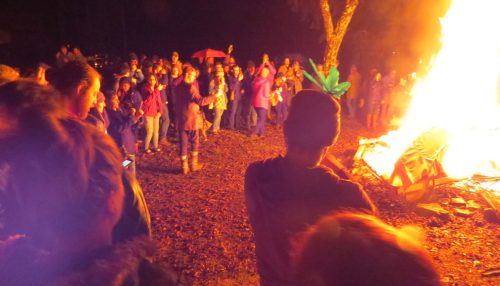 A crowd gathers around a large bonfire at night, with people enjoying the warmth and atmosphere.