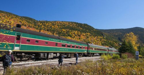 A colorful train on the Conway Scenic Railroad surrounded by vibrant autumn foliage and clear blue skies.