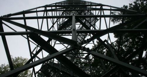 A low-angle view of a tall metal tower surrounded by trees, with a cloudy sky in the background.