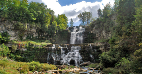 A scenic waterfall cascading down rocky cliffs, surrounded by lush greenery and a blue sky with fluffy clouds.