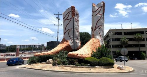 Giant cowboy boots sculpture in a roundabout, surrounded by greenery and a blue car parked nearby.