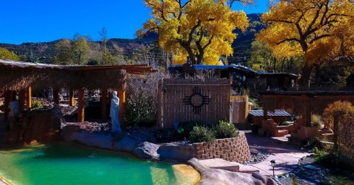 A serene outdoor spa with a green pool, surrounded by autumn trees and rustic wooden structures under a clear blue sky.