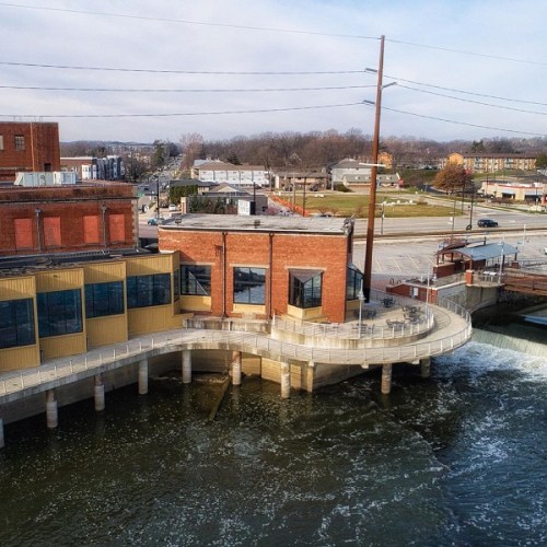 Aerial view of a brick building by a river, featuring a curved walkway and flowing water, with a cityscape in the background.