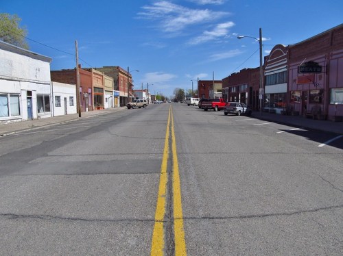 A wide, empty street lined with buildings and parked cars under a clear blue sky.