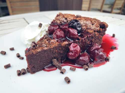 A slice of chocolate cake topped with cherries, chocolate shavings, and a dollop of whipped cream on a white plate.