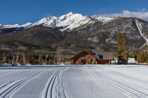 A snowy landscape with groomed ski trails, a wooden lodge, and majestic mountains in the background under a clear blue sky.