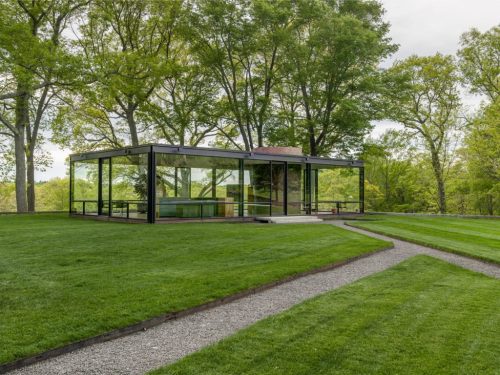A modern glass house surrounded by lush greenery and a manicured lawn, with a gravel pathway leading to it.