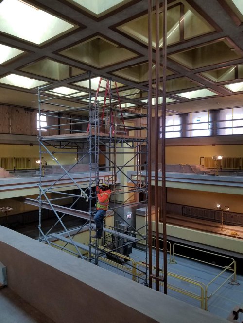 A construction worker on a scaffold inside a building with a coffered ceiling and yellow walls.