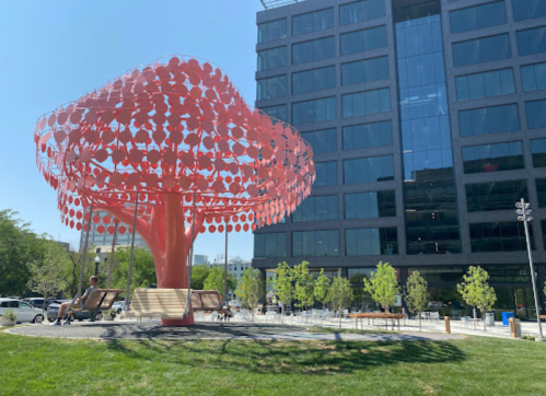 A large, vibrant red tree sculpture stands in a park next to a modern building, with benches and greenery around.