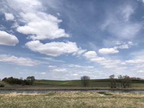 A serene landscape featuring a calm river, grassy banks, and a blue sky dotted with fluffy white clouds.