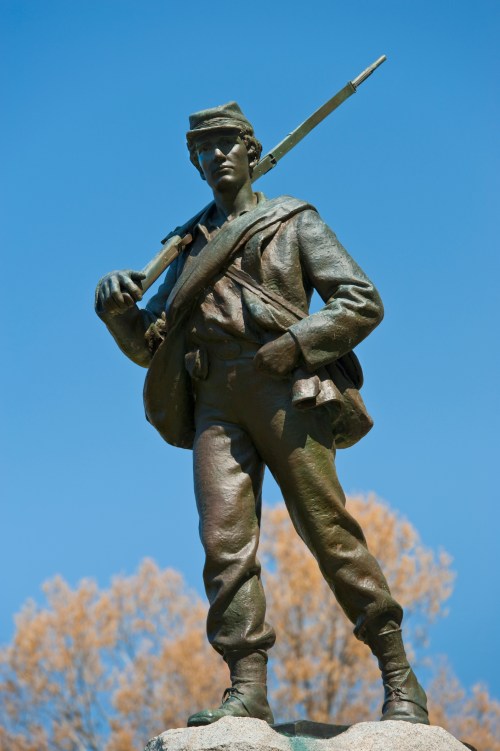 A bronze statue of a young soldier holding a rifle, standing confidently against a clear blue sky.