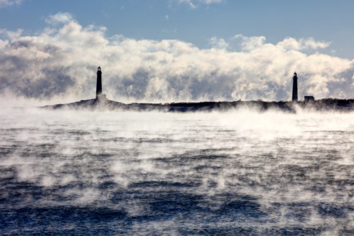 Two lighthouses stand in the distance, surrounded by mist rising from a cold, choppy sea under a cloudy sky.