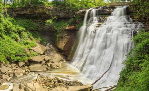 A serene waterfall cascades over rocky cliffs, surrounded by lush greenery and flowing water below.