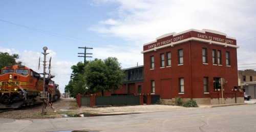 A red brick freight depot building with a train passing by, set against a cloudy sky.