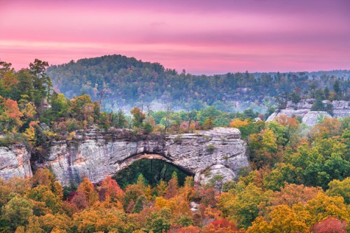 A scenic view of a natural stone arch surrounded by vibrant autumn foliage and a colorful sky at sunset.