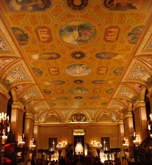 Ornate ceiling with intricate designs and paintings, illuminated by chandeliers in a grand hall.