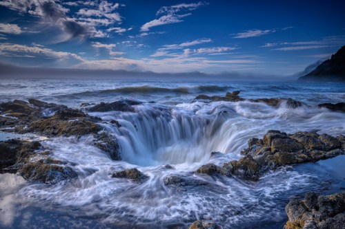 A dramatic seascape featuring swirling waves crashing over rocky formations under a blue sky.