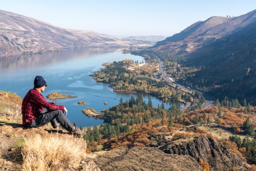 A person sits on a rocky ledge overlooking a river valley surrounded by autumn-colored trees and mountains.