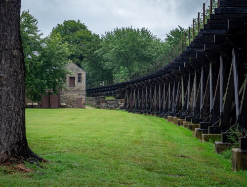 A grassy area with a stone building and a long, elevated structure supported by metal beams in a wooded setting.
