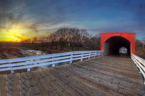 A red covered bridge at sunset, with a wooden walkway and trees in the background, reflecting a serene landscape.