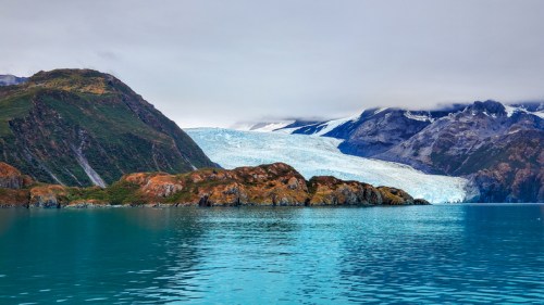 A serene landscape featuring a glacier flowing down mountains into a turquoise body of water, surrounded by lush greenery.