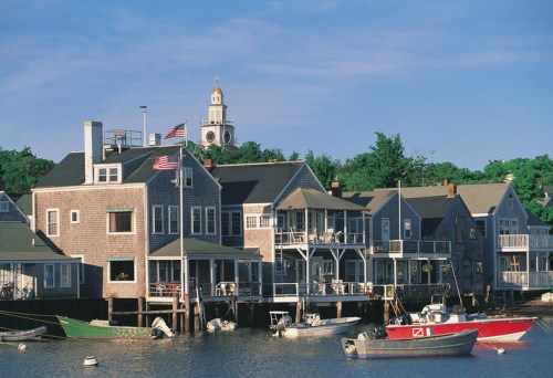 Quaint waterfront houses with boats, flags, and a church steeple in the background under a clear blue sky.