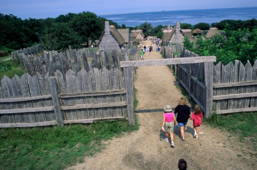 A family walks through a wooden gate towards a historic village by the ocean, surrounded by greenery and thatched-roof huts.