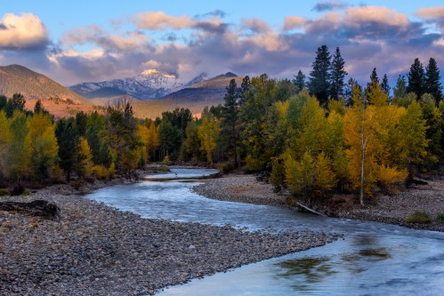 A winding river flows through a forest of autumn trees, with snow-capped mountains and clouds in the background.