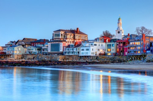Colorful buildings line a waterfront at dusk, reflecting in calm water with a church steeple in the background.