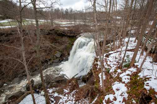 A waterfall cascades over rocky terrain, surrounded by bare trees and patches of snow in a serene landscape.